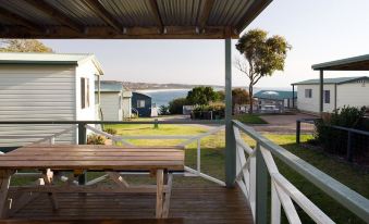 a wooden deck with a picnic table and chairs , overlooking a grassy area and water at NRMA Merimbula Beach Holiday Resort