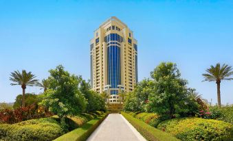 a tall , modern building with blue glass windows and a glass entrance is surrounded by green trees and bushes at The Ritz-Carlton, Doha