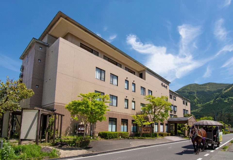 a modern hotel building with multiple floors , surrounded by trees and mountains , under a clear blue sky at Yufuin Hotel Shuhokan