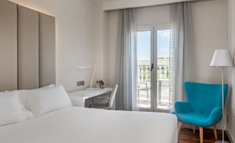 a hotel room with a white bed , a blue chair , and a window overlooking the city at NH Madrid Nacional