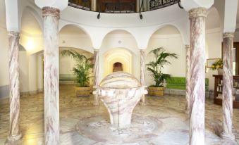 a large , white marble fountain with a palm tree in the center is surrounded by potted plants at Le Grand Hotel