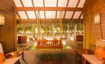a wooden deck with a bench , chairs , and tables under a thatched roof overlooking the beach at Medhufushi Island Resort
