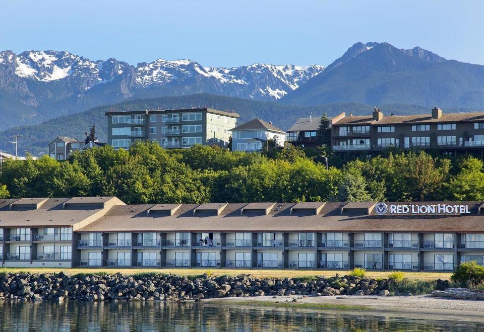 a large building with a red logo is situated on the shore of a lake , with mountains in the background at Red Lion Hotel Port Angeles Harbor