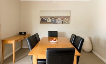 a dining table with black chairs and a bowl of fruit in the center , set against a beige wall with a painting of a row of houses at The Marina Hotel - Mindarie