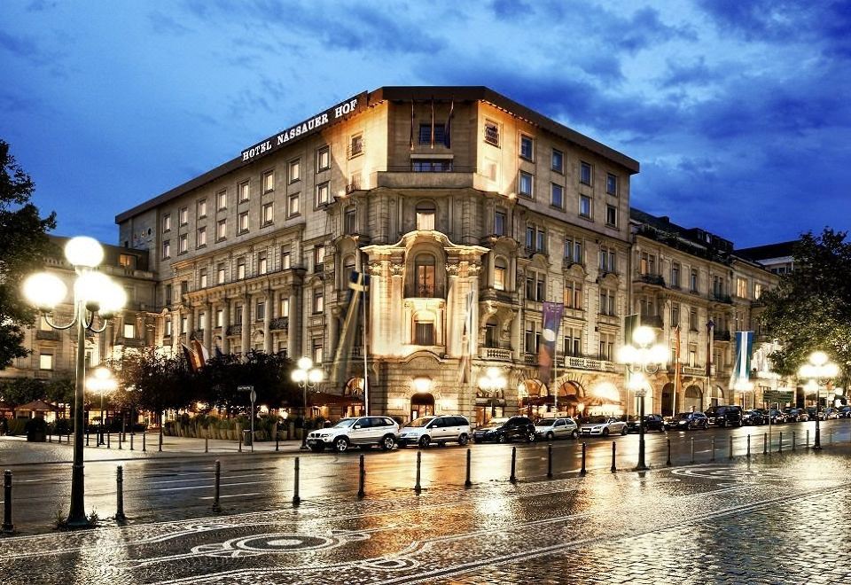 a large , ornate building with a well - lit facade is situated on a wet street near parked cars and streetlights at Hotel Nassauer Hof