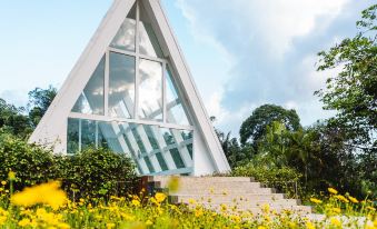 a modern , triangular - shaped house with large windows and a sloping roof , surrounded by lush greenery and yellow flowers at Horison Green Forest Bandung