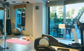 a well - equipped gym with various exercise equipment , including a treadmill and weights , positioned near large windows that offer views of outdoor dining areas at Moxy Patra Marina