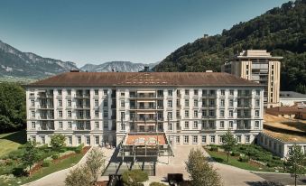 a large white building with a brown roof and balconies is surrounded by trees and mountains at Grand Resort Bad Ragaz