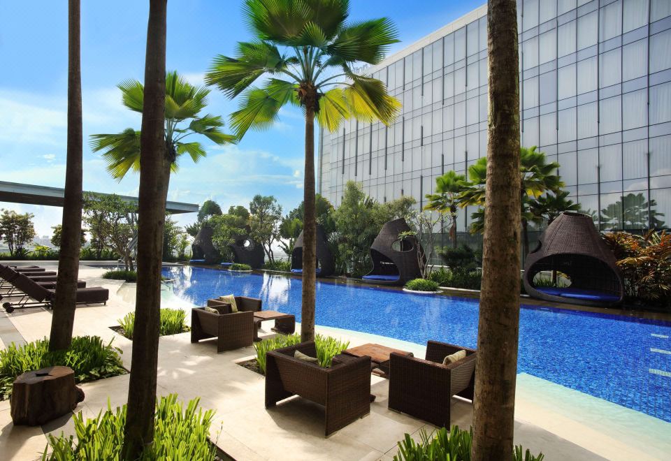 a large outdoor pool surrounded by palm trees , with several lounge chairs and tables placed around the pool area at Hilton Bandung