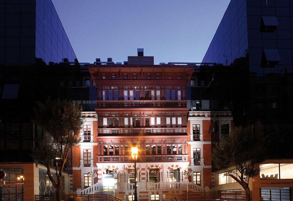 a city street at night with a red brick building illuminated by lights , and a couple walking down the sidewalk at Barcelo Hotel Oviedo Cervantes