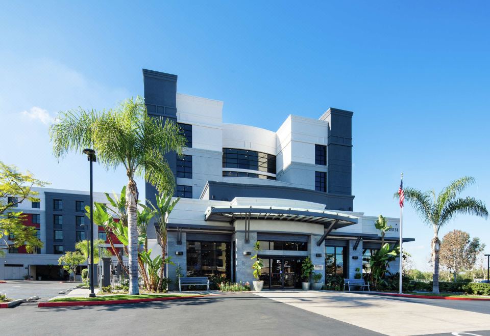 a modern hotel building with a large entrance , surrounded by palm trees and clear blue skies at Hilton Garden Inn Irvine Spectrum Lake Forest