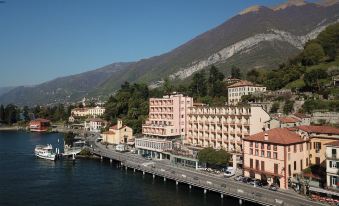 a large pink building is situated on a waterfront with mountains in the background , and boats floating on the water at Hotel Bazzoni