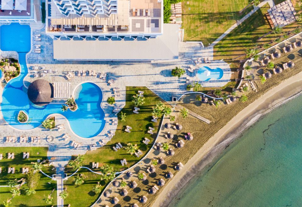 aerial view of a resort with a large pool surrounded by lounge chairs and umbrellas , as well as a grassy area near the beach at Golden Bay Beach Hotel