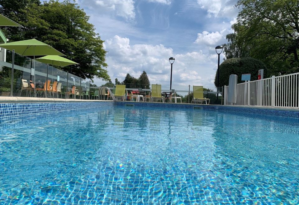 a large outdoor swimming pool surrounded by a fence , with lounge chairs and umbrellas placed around the pool area at Holiday Inn Lille - Ouest Englos