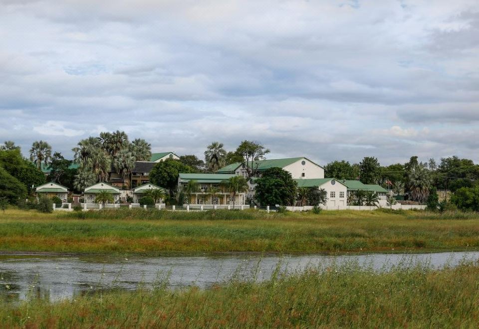 a grassy field with a small pond in the middle , surrounded by several buildings on the other side at Maun Lodge