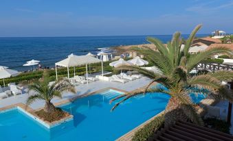 a large swimming pool with a palm tree and lounge chairs is surrounded by white umbrellas and a beach house at Aldemar Knossos Royal