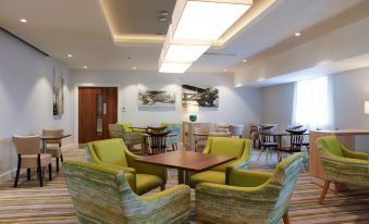 a modern lounge area with green chairs and a wooden table in the center , surrounded by white walls and blue ceiling at Hampton by Hilton Exeter Airport