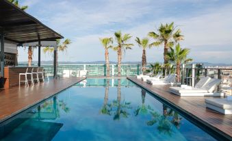 a large , empty swimming pool is surrounded by palm trees and a wooden deck , with a view of the ocean in the background at Renaissance Barcelona Fira Hotel