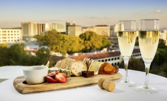 a table with a plate of food and two glasses of wine in front of a cityscape at Novotel Canberra