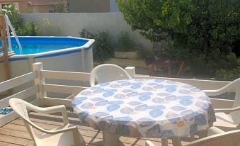 a wooden deck with a blue and white tablecloth covering a dining table , surrounded by white chairs and a swimming pool in the background at Les Amandiers