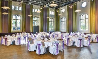 a large , empty banquet hall with purple and green draping , white chairs , and multiple tables set for a formal event at Steigenberger Braunschweig