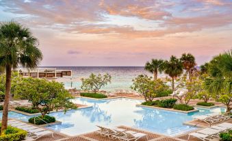 a beautiful swimming pool area with a pool , lounge chairs , and trees , overlooking the ocean at sunset at Curacao Marriott Beach Resort