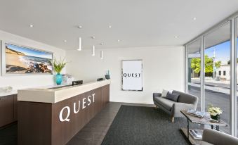 "a reception area with a wooden counter , couch , and potted plant , along with a sign that reads "" quest ""." at Quest Cheltenham