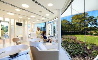 a modern white room with a large window overlooking a green lawn and trees , and a person sitting in a chair at Racv Royal Pines Resort Gold Coast