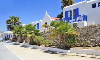 a white house with blue windows and a stone wall , surrounded by palm trees and plants at Mykonos Beach Hotel