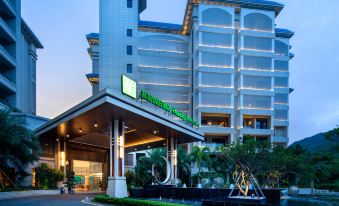 The front entrance of a hotel is depicted, featuring an illuminated sign above it, with other buildings in the background at Holiday Inn & Suites Sanya Yalong Bay