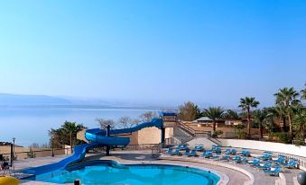 a large blue pool with a water slide is surrounded by lounge chairs and palm trees at Dead Sea Spa Hotel