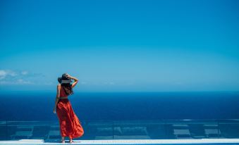 a woman in a red dress and straw hat stands on the edge of a pool with a blue ocean and blue sky in the background at Emerald Villas & Suites - the Finest Hotels of the World