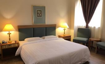 a large bed with white sheets and a wooden headboard is in a room with two chairs , a window , and a painting at The Executive Hotel Lahad Datu