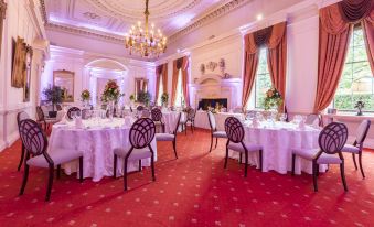 a large dining room with multiple tables set up for a formal event , surrounded by chairs and lit by hanging chandeliers at Coombe Abbey Hotel