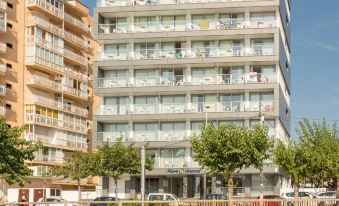 a tall , white building with many windows is situated next to a smaller one , and trees and a parking lot in front of it at Pierre & Vacances Blanes Playa