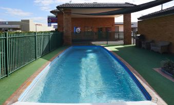 a long , narrow swimming pool is surrounded by a green fence and a brick building at Adelong Motel