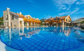 a large , modern swimming pool with a blue tile border is surrounded by wooden houses and has umbrellas placed around it at Dalyan Live Spa Hotel
