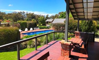 a wooden deck with a bench , surrounded by trees and a swimming pool , under a clear blue sky at Prom Coast Holiday Lodge