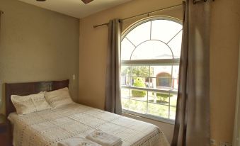 a cozy bedroom with a large window , white bed , and beige curtains , under a ceiling fan at Villa Del Sol