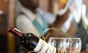 a man is standing behind a bar , holding a wine bottle and two glasses of wine at Maun Lodge