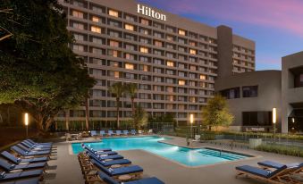 an outdoor pool surrounded by lounge chairs and umbrellas , with a hilton hotel in the background at Hilton Los Angeles-Culver City, CA