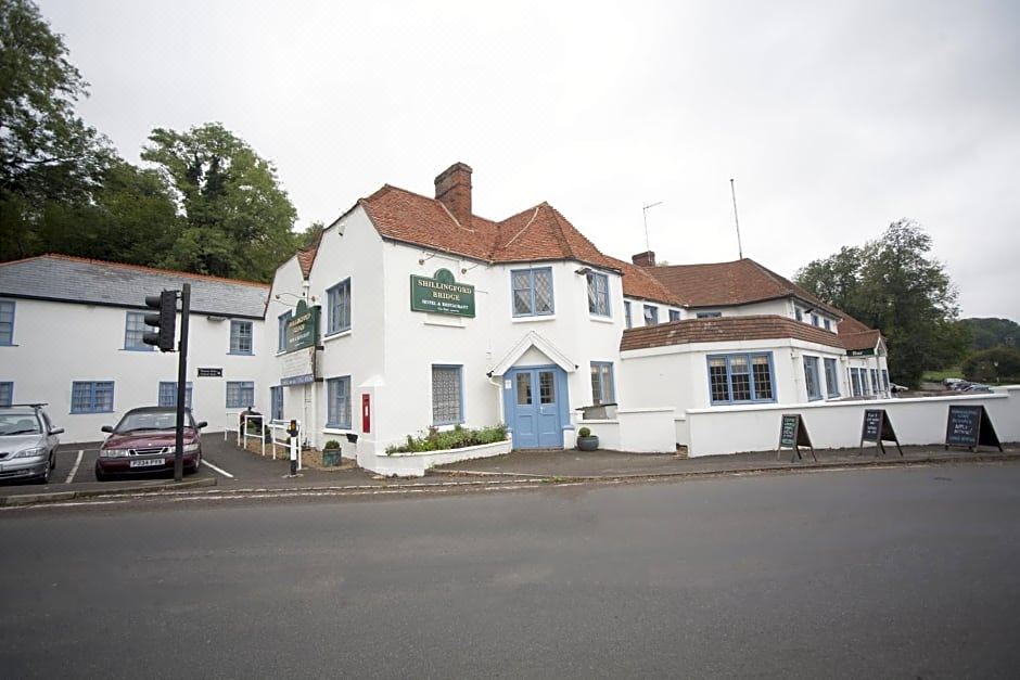 a white building with a red roof and green sign , situated on a street corner in front of trees at Shillingford Bridge Hotel