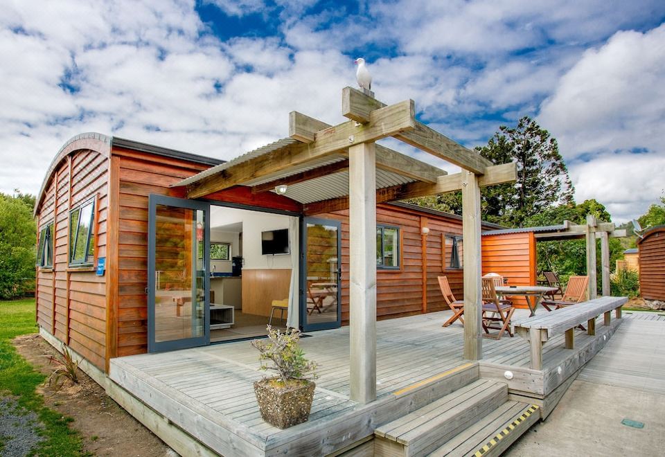 a modern wooden house with a covered porch and outdoor furniture , under a blue sky with clouds at Hot Water Beach Top 10 Holiday Park