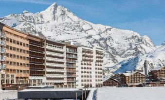 a snow - covered mountain with a hotel in the background , surrounded by buildings and snow - covered trees at Langley Hotel Tignes 2100