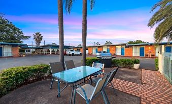 a glass dining table with chairs is set up in an outdoor patio area with palm trees and parked cars at Plainsman Motel