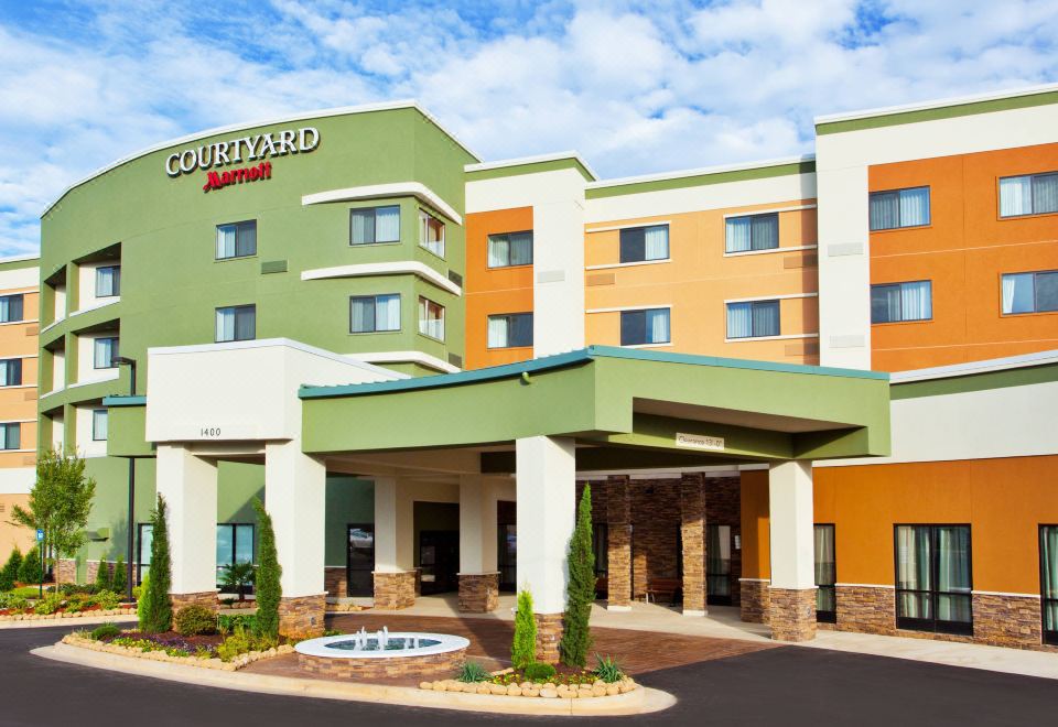 a courtyard by marriott hotel with a green and orange facade , a fountain in the foreground , and palm trees surrounding it at Courtyard Columbus Phenix City/Riverfront