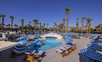 a large swimming pool surrounded by lounge chairs and umbrellas , with palm trees in the background at GrandResort by Leonardo Hotels