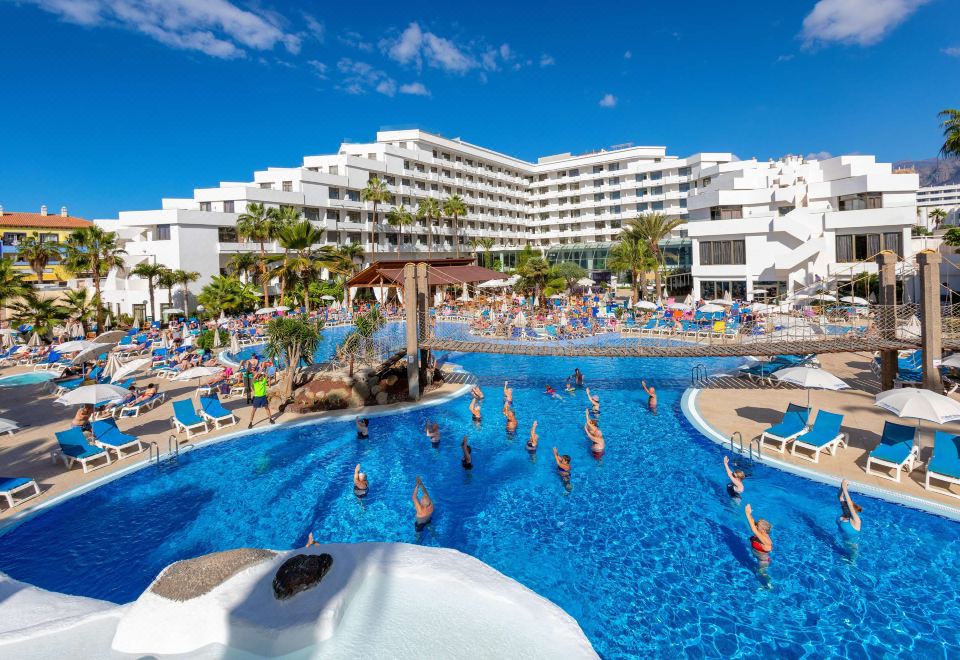 a large , crowded swimming pool with people in the water and on the beach , surrounded by white buildings at Hotel Best Tenerife