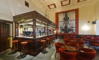 a bar with wooden stools and red chairs , a wooden counter , and a large picture of a ship on the wall at Royal Maritime Hotel