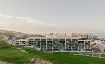 a large , modern building with a parking lot in front of it , surrounded by green grass and mountains in the background at Nobu Hotel Los Cabos